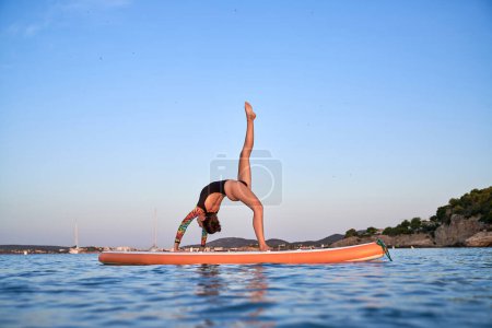 Téléchargez les photos : Vue de côté pleine longueur de femme mince en maillot de bain debout dans la pose de roue avec jambe levée sur SUP board en mer bleue avec paysage à couper le souffle - en image libre de droit