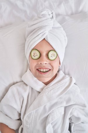 Photo for Smiling child with cucumber slices on eyes wearing bathrobe and towel turban relaxing on soft bed during beauty procedures in bedroom at home - Royalty Free Image