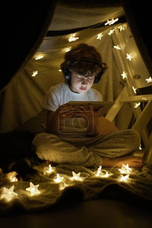 Photo for Full body of serious boy in headphones watching video on modern tablet while sitting with crossed legs near glowing garland in dark room - Royalty Free Image