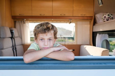 Téléchargez les photos : Garçon détendu avec les bras croisés regardant la caméra tout en se reposant derrière la fenêtre de la caravane moderne pendant les vacances d'été - en image libre de droit