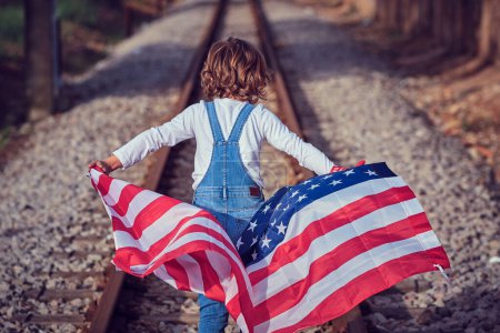 Téléchargez les photos : Vue arrière d'un enfant méconnaissable en denim marchant sur le chemin de fer avec le drapeau de l'Amérique par une journée ensoleillée - en image libre de droit
