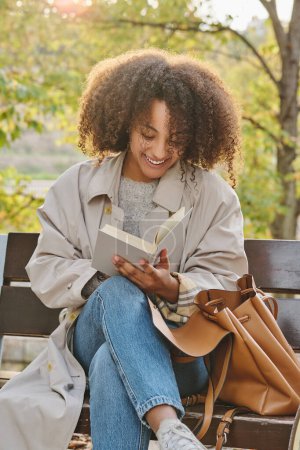 Photo for Positive young African American female student in casual outfit sitting on wooden bench in park and reading book while preparing for exam - Royalty Free Image