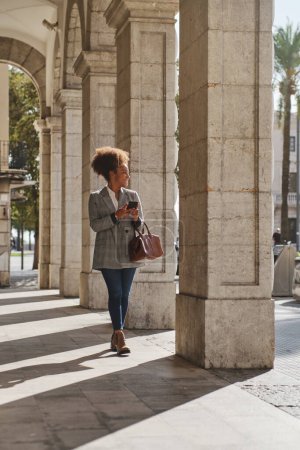 Téléchargez les photos : Contenu ethnique femelle avec sac à main marchant sur le trottoir près des colonnes de pierre à la lumière du soleil et regardant loin tout en envoyant des textos sur téléphone portable - en image libre de droit
