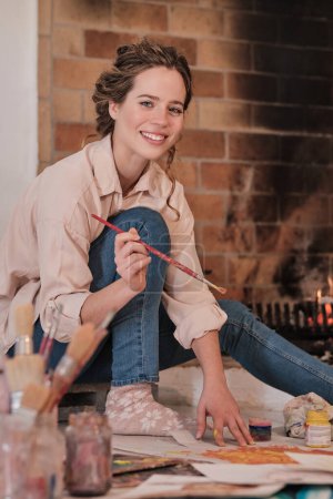 Téléchargez les photos : Vue latérale de l'artiste féminine positive en vêtements décontractés assis sur le sol près d'une cheminée en feu et regardant la caméra tout en peignant image - en image libre de droit