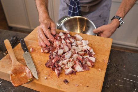 Photo for Unrecognizable male chef in apron standing at kitchen counter with chopping board and knife while cooking at home and pouring octopus into metal bowl - Royalty Free Image