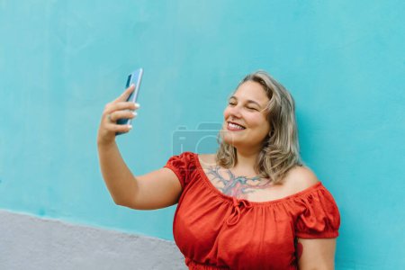 Foto de Ángulo alto de mujer joven de talla grande en ropa casual tomando selfie con teléfono inteligente mientras está de pie sobre fondo de pared azul al aire libre - Imagen libre de derechos