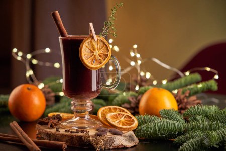 Téléchargez les photos : Verre de vin chaud alcoolisé servi sur la table avec des oranges et des branches de conifères lors de la célébration de Noël dans une chambre avec guirlande lumineuse - en image libre de droit