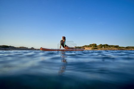 Téléchargez les photos : Athlète féminine flexible en maillot de bain pratiquant le yoga sur paddleboard et prenant la pose de Bhujangasana parmi l'eau de mer ondulante - en image libre de droit