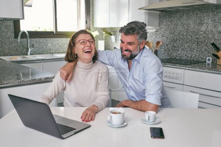Foto de Hombre maduro sonriente abrazando a la mujer mayor mientras está sentado en la mesa blanca con tazas de té y viendo videos divertidos en la computadora portátil en la cocina moderna en casa - Imagen libre de derechos
