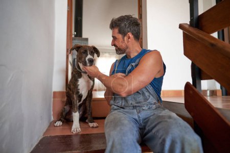 Téléchargez les photos : Aimant propriétaire masculin en denim chien caressant ensemble dans les escaliers à la maison - en image libre de droit