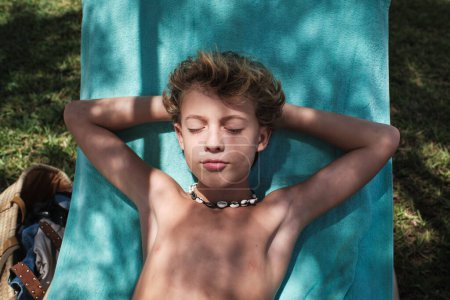 Photo for From above of shirtless boy with blond hair lying on blue blanket with hands clasped behind head while relaxing in garden with eyes closed - Royalty Free Image