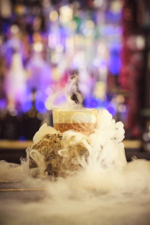 Foto de Cóctel creativo caliente en soporte de piedra en medio del vapor servido en el mostrador en el moderno bar por la noche - Imagen libre de derechos