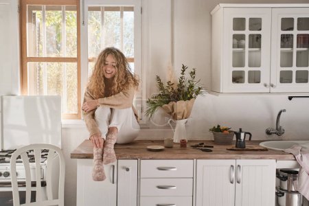 Foto de Cuerpo completo de mujer joven en ropa casual sentada en los muebles de la cocina y sonriendo en la acogedora cocina en casa - Imagen libre de derechos