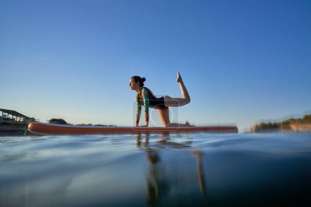 Téléchargez les photos : Pleine longueur d'athlète féminine sportive avec les yeux fermés prenant la pose de Vyaghrasana sur le tableau SUP tout en pratiquant le yoga parmi la mer ondulante en soirée d'été - en image libre de droit