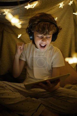 Foto de Niño feliz en auriculares con auriculares viendo interesante video en la tableta moderna mientras está sentado cerca de la guirnalda brillante en la habitación acogedora - Imagen libre de derechos