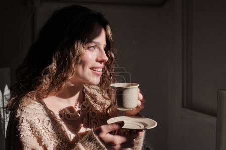 Téléchargez les photos : Vue latérale de la jeune femme avec tasse de café chaud souriant et regardant loin tout en se reposant dans une chambre confortable à la maison - en image libre de droit