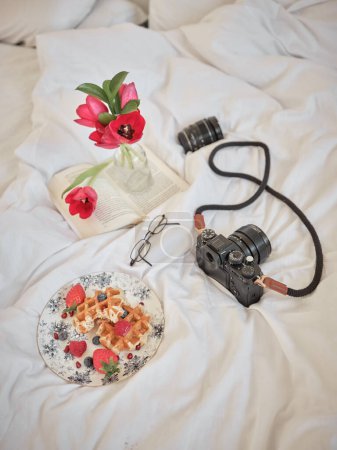 Téléchargez les photos : Assiette avec gaufres sucrées et tasse à thé sur lit blanc avec smartphone et appareil photo près du bouquet de tulipes dans la chambre - en image libre de droit