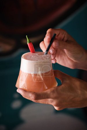 Téléchargez les photos : Crop person démontrant délicieux cocktail décoré avec du piment et servi dans un verre avec de la paille sur fond flou - en image libre de droit