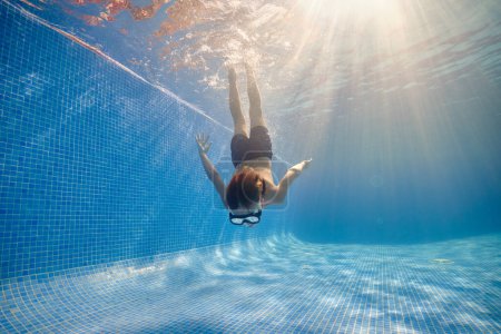 Foto de Cuerpo completo de chico descalzo en gafas y pantalones cortos nadando en piscina limpia bajo el sol - Imagen libre de derechos
