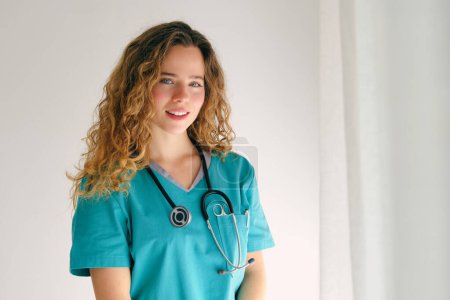 Téléchargez les photos : Jeune femme médecin positive avec de longs cheveux bouclés portant l'uniforme médical et stéthoscope debout près du mur blanc et regardant la caméra - en image libre de droit