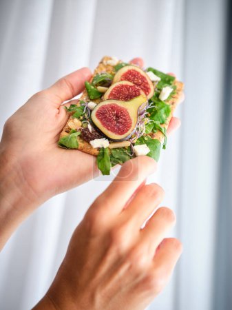 Photo for Unrecognizable person showing fresh healthy fig slices decorated with mint leaves and salad on bread while touching white cheese piece with finger against white curtain background - Royalty Free Image