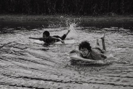 Foto de Blanco y negro de niños varones alegres en uniforme escolar nadando con tabla mientras salpica y crea olas en el agua a la luz del día - Imagen libre de derechos