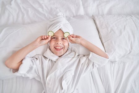 Foto de Niño feliz con turbante de toalla y albornoz sonriendo y mirando a la cámara mientras se relaja en la cama y se quita la máscara de pepino de los ojos en el dormitorio en casa - Imagen libre de derechos