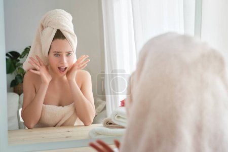 Photo for Astonished female wrapped in towels looking at reflection of mirror with opened mouth at table during daily routine in light room - Royalty Free Image