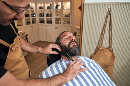 Téléchargez les photos : Cultiver barbier en uniforme barbe de coupe du client avec des ciseaux tout en travaillant dans le salon de coiffure - en image libre de droit