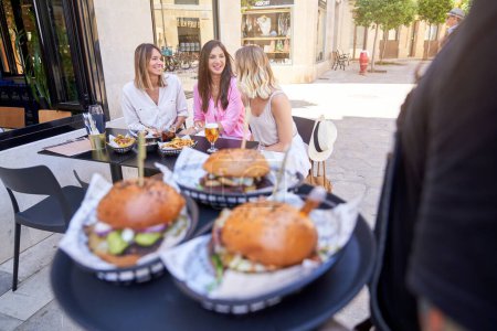 Téléchargez les photos : Crop man plateau de service avec des hamburgers pour les clientes souriantes assis à table et parlant entre eux dans le café de trottoir - en image libre de droit