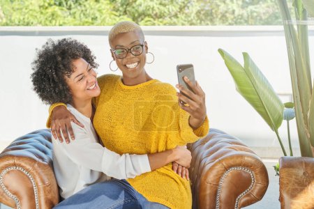 Photo for Smiling multiracial homosexual females in casual clothes having fun while sitting in armchair and taking self portrait on mobile phone together - Royalty Free Image