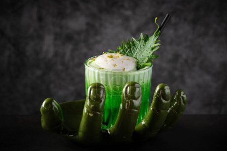 Téléchargez les photos : Verre de cocktail vert froid rafraîchissant décoré de feuilles de shiso et servi sur une assiette en forme de main placée sur le comptoir dans une pièce sombre - en image libre de droit