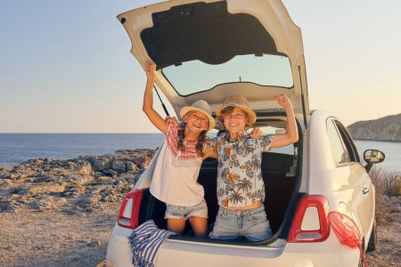 Téléchargez les photos : Deux enfants se penchant dans le coffre de la voiture les mains levées dans un geste de victoire en faisant face à la caméra et portant un chapeau de paille avec des paniers d'objets pour la plage à côté de la voiture avec la mer en arrière-plan - en image libre de droit