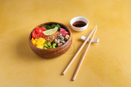 Photo for High angle of wooden bowl with slices of healthy tomato mango tofu and radish served with wakame salad and edamame beans with piece of lime on top near chopsticks and soy sauce in gravy boat on yellow table - Royalty Free Image