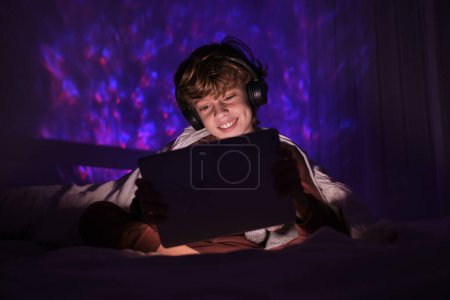 Téléchargez les photos : Enfant joyeux dans les écouteurs tablette de navigation tout en étant assis sur le lit sous la couverture dans la chambre à coucher avec une faible lumière la nuit - en image libre de droit