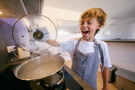 Photo for Positive child in apron with lid against boiling water in saucepan on stove during cooking process at home - Royalty Free Image