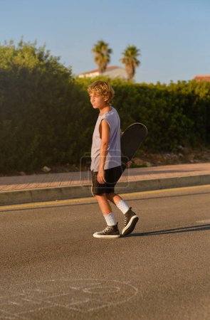 Foto de Vista lateral de niño preadolescente tranquilo en ropa hipster y con monopatín caminando por la carretera en la noche de verano y mirando hacia otro lado - Imagen libre de derechos