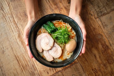 Téléchargez les photos : Vue du dessus d'une personne anonyme tenant une assiette noire avec une soupe japonaise fraîche et cuite à base de bouillon crémeux porc et nouilles sur une table en bois - en image libre de droit