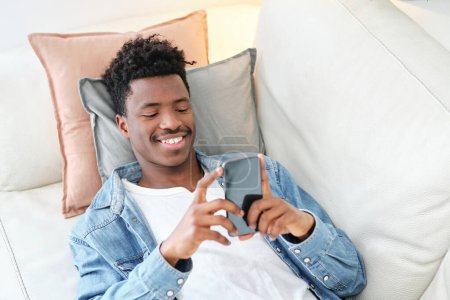 Photo for High angle smiling African American male in denim jacket browsing modern mobile phone and lying on comfortable sofa - Royalty Free Image