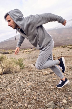 Foto de Vista lateral completa de atleta masculino hispano concentrado en ropa deportiva gris cálida corriendo rápido en terreno pedregoso cerca del Teide de montaña en Tenerife Islas Canarias España - Imagen libre de derechos