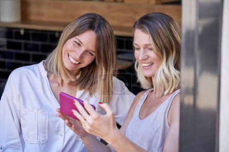 Téléchargez les photos : Positive jeune femme souriant tout en montrant des photos sur téléphone portable à petite amie pendant le week-end dans le café - en image libre de droit