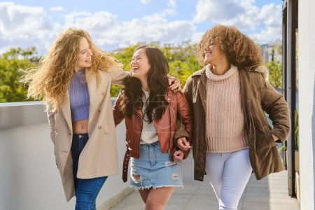 Foto de Alegre joven diversas novias riendo de broma y caminando en la terraza en ropa de abrigo en otoño mientras se miran entre sí - Imagen libre de derechos
