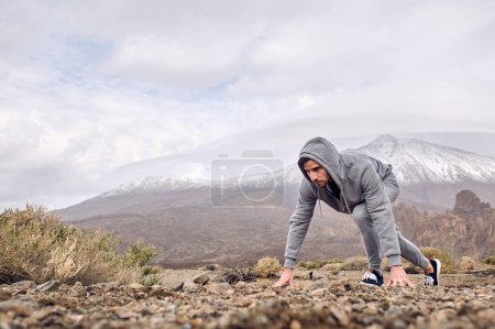 Téléchargez les photos : Vue de côté corps entier du voyageur hispanique concentré masculin en vêtements de sport debout en position de départ accroupi sur un terrain pierreux contre la montagne Teide à Tenerife Îles Canaries Espagne - en image libre de droit