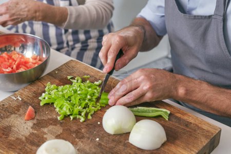 Photo for Crop faceless male cutting fresh green pepper with knife on cutting board with onion while cooking near woman in light kitchen - Royalty Free Image