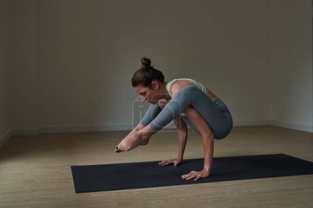 Téléchargez les photos : Vue latérale d'une athlète féminine concentrée en tenue de sport debout sur un tapis noir dans la pose de Tittibhasana et regardant vers le bas pendant l'entraînement de yoga dans un studio spacieux - en image libre de droit
