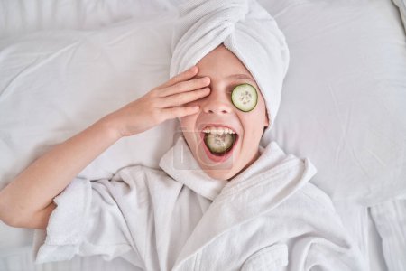 Photo for Playful boy in towel turban and bathrobe eating cucumber slice and covering eye with hand while resting on white bed in bedroom at home - Royalty Free Image