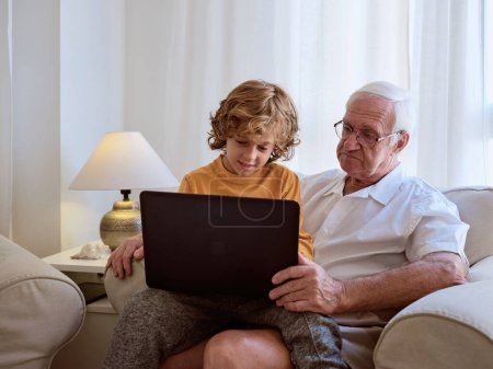 Téléchargez les photos : Petit-fils mignon naviguant netbook moderne tout en étant assis sur les tours de grand-père dans les lunettes dans le fauteuil dans le salon à la maison - en image libre de droit