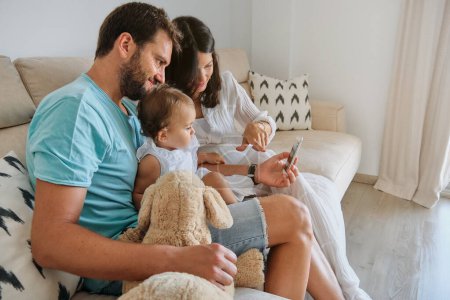 Photo for Side view of family in casual clothes sitting on sofa and watching video on smartphone while spending time together at home - Royalty Free Image