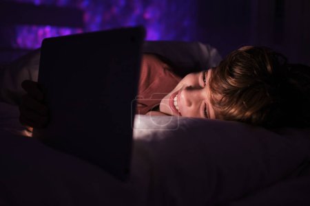 Téléchargez les photos : Garçon ravi de regarder la vidéo intéressante sur tablette moderne tout en étant couché dans le lit dans la chambre avec une lumière tamisée la nuit - en image libre de droit