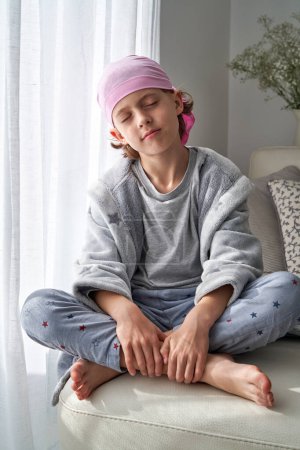 Photo for Full body of barefoot serene kid sitting with closed eyes and legs crossed in pink headscarf for participating in movement of breast cancer awareness siting on couch - Royalty Free Image